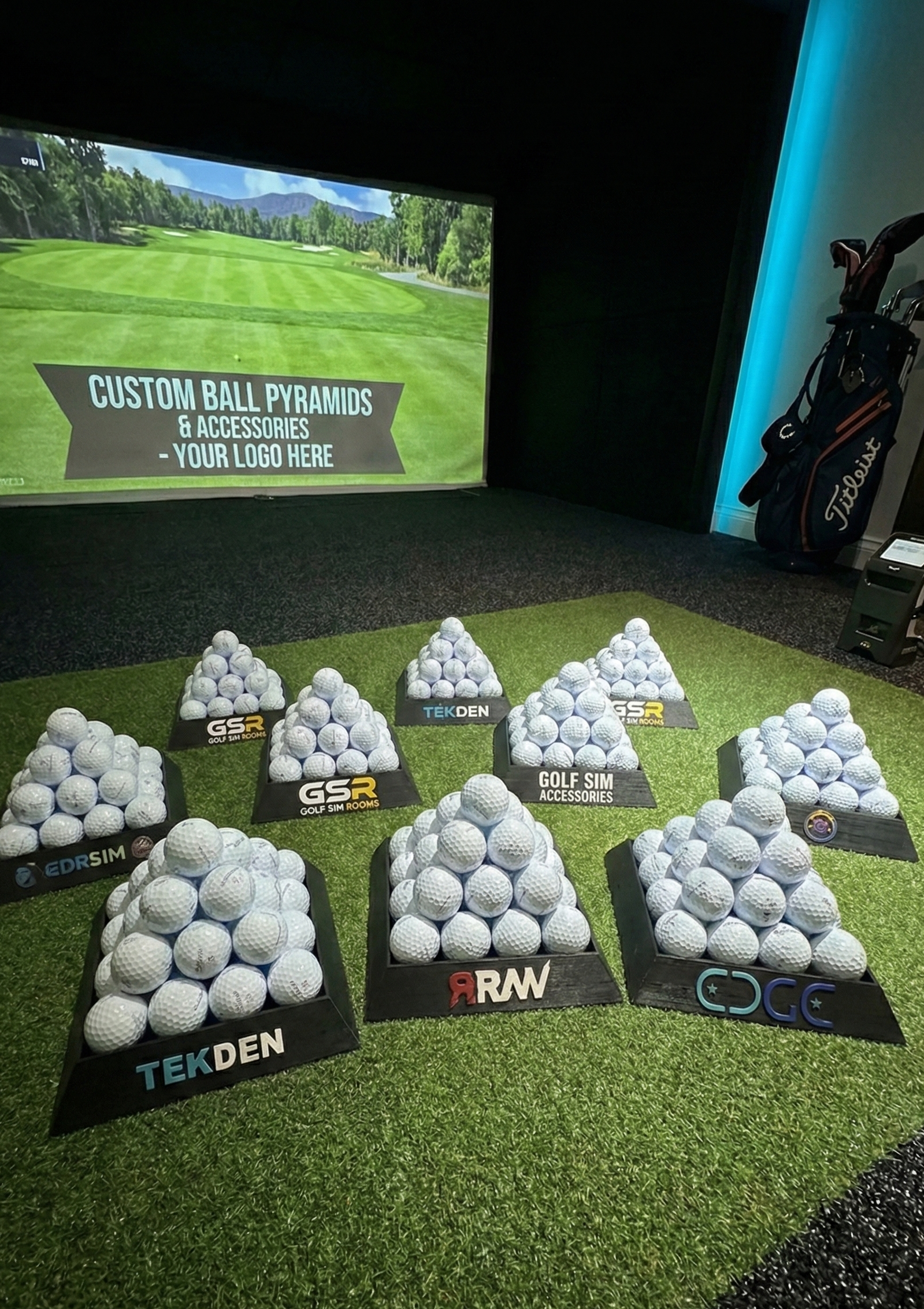 Golf ball pyramids with various brands displayed on a grass-like surface in front of a screen showing a golf course.